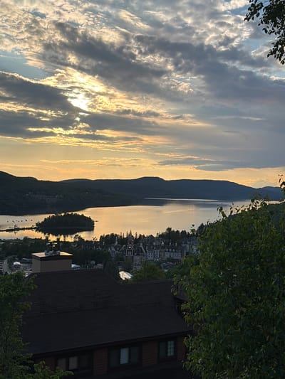 This is an outdoor, high-angle, full shot of a resort village at dusk, situated in Mont-Tremblant, Canada. The sky is filled with dramatic, patchy clouds illuminated by the setting sun, casting a warm glow over the scene. In the foreground, the dark, sloped roof of a building is visible, with some chimneys protruding from it. To the right, lush green foliage partially obscures the view.
In the midground, the village of Mont-Tremblant unfolds. Numerous buildings with red roofs and architectural details are nestled together on a slope leading down to a large lake. The lake itself reflects the sky and the surrounding hills, creating a shimmering surface. A small, tree-covered island sits in the middle of the lake.
The background is dominated by rolling, tree-covered hills that form the natural landscape surrounding the resort. The overall impression is one of tranquility and natural beauty, typical of a mountain resort town during sunset. There are no people visible in the image.