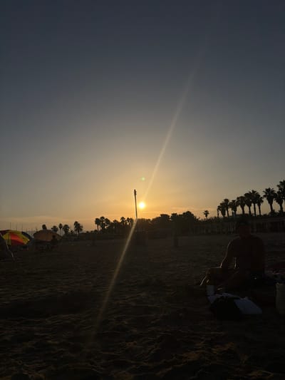 The image captures a sunset scene on a beach, likely in Salou, Spain, given the presence of palm trees lining the background. The sky is a gradient of pale yellow to a deep blue, with the sun positioned low on the horizon, casting a bright, lens-flare effect diagonally across the frame. In the foreground, the sandy beach is textured with ripples. To the right, a person is seated on the sand, silhouetted against the bright sky, wearing swim trunks and appearing to be relaxed. Nearby, a few items are placed on the sand, including what appears to be a towel and a dark bag. In the mid-ground, several beach umbrellas, some colorful and others a muted orange, provide shade. Behind them, a line of palm trees and some low-lying structures are visible, also silhouetted. The overall atmosphere is tranquil, typical of a beach at dusk.