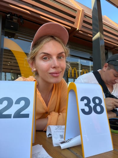 In Sedaví, Spain, a medium shot captures a blonde woman wearing a pink baseball cap and an orange top, seated at an outdoor table. She is looking towards the camera with a slight smile, her face well-lit. To her left, a white table marker displays the number "22." To her right, another white table marker shows the number "38." In the background, the exterior of a McDonald's restaurant is visible, identifiable by the iconic golden arches. A person wearing a black cap and white shirt is seated to the right of the woman, looking down at a mobile phone. The overall scene suggests a casual dining experience outdoors at a McDonald's.