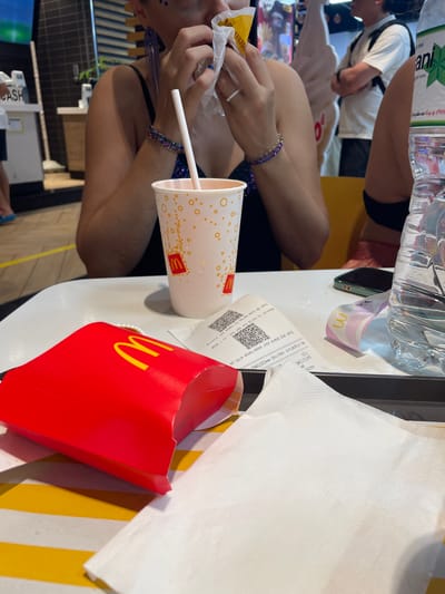 The image captures a close-up view of a table setting at a McDonald's restaurant in Thuận An, Vietnam. The focus is on a red McDonald's french fry container, an open white paper wrapper with food inside being held by a person, a white paper cup with the McDonald's logo and orange circular pattern, a plastic straw, a receipt with QR codes, a phone with a green case, and a partially visible plastic water bottle. In the background, blurred figures of people and restaurant decor are visible, including a counter with a screen that displays "CASH". The overall scene suggests a meal in progress or just concluded, with the items arranged on a white table surface.