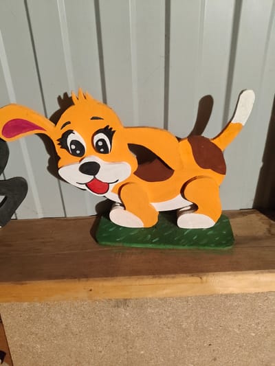 This is a close-up, eye-level, medium shot of a wooden, cartoon-like dog figurine. The figurine is predominantly orange with brown patches on its back, white paws and muzzle, and pink inner ears. It has large, black and white eyes with eyelashes, black eyebrows, a black nose, and a red tongue sticking out. The dog is standing on a green, rectangular base that resembles grass. The figurine is placed on a wooden shelf, and behind it is a corrugated metal wall. To the left of the dog, a portion of a black object is visible, which appears to be another figurine or decoration. The lighting casts shadows on the wall behind the dog. The scene is indoors, likely in a workshop or storage area, given the materials and unfinished look of the background and shelf. The specific location in Staroye Drozhzhanoye, Russia, cannot be determined from the visual information provided in the image.