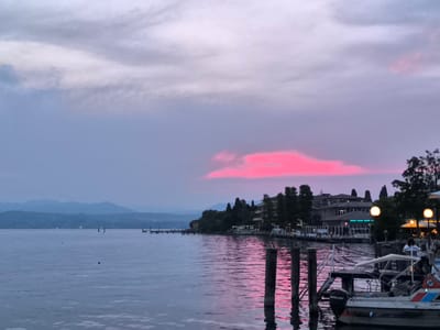 The image captures a serene sunset scene on a lake, likely Lake Garda, given the context of Sirmione, Italy. The sky is painted with soft hues of purple and pink, with a particularly vibrant streak of fuchsia cloud in the middle distance, suggesting a dramatic end to the day. The water is calm and reflects the colors of the sky, creating a shimmering, ethereal effect.

Along the shore, a picturesque town is visible, characterized by buildings with terracotta roofs and a line of trees. Several boats are docked at a pier, some with awnings, indicating a popular waterfront area. The lighting from the town and some of the boats suggests that evening has begun to settle in, with artificial lights starting to illuminate the scene.

In the foreground, two wooden posts rise from the water, their reflections elongated in the glassy surface. A small boat is moored to a dock, with the visible text "MERCURY" on its outboard motor. Two people are visible near the boat, one standing and the other seated, though their features are not distinct. Further down the pier, more figures can be seen, enjoying the lakeside atmosphere.

In the far distance, a mountain range forms a hazy silhouette against the fading light. The overall mood of the image is peaceful and tranquil, evoking a sense of calm and beauty associated with a lakeside sunset. The weather appears clear, with the dramatic sky being the primary atmospheric element.