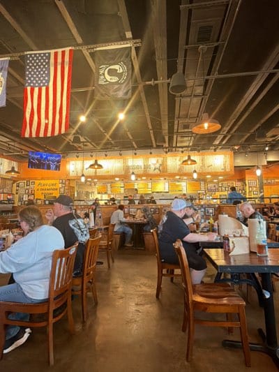 The image depicts the interior of a Mission BBQ restaurant, a chain that is located in Rochester, New York. The space is filled with wooden tables and chairs occupied by patrons. Banners and flags hang from the ceiling alongside lighting fixtures. A counter at the back serves as the order and pickup area. The room is decorated with what appears to be memorabilia. The floor is concrete.
