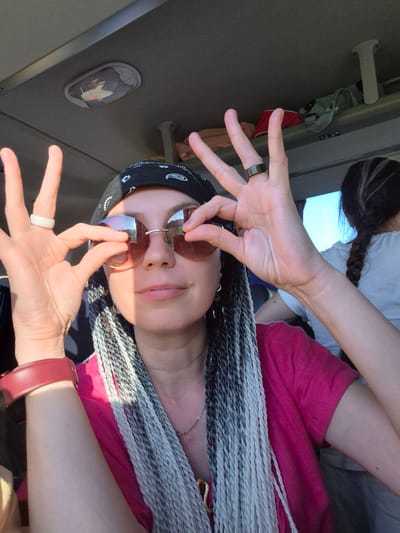A selfie taken inside a vehicle, likely a bus, shows a woman with braided hair and sunglasses. She is wearing a pink shirt and a black bandana. Her hands are raised, and she is making a peace sign with her right hand, while her left hand is near her sunglasses. The interior of the vehicle is visible in the background, with luggage on a shelf. The lighting suggests it is daytime. The location is the city of Verkhniye Tatyshly, Russia.