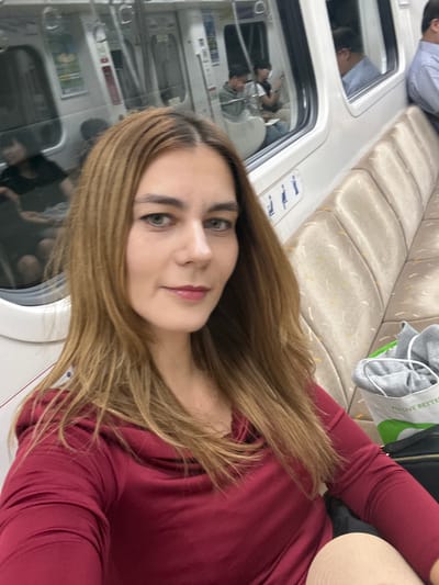 A woman wearing a maroon long-sleeved shirt is on a train, likely in Gimpo-si, South Korea. She is seated next to a window, with a beige upholstered bench behind her. Other passengers are visible through the train windows. A gray bag with green and white lettering is on the bench next to her. The image is taken from a close-up, selfie perspective.