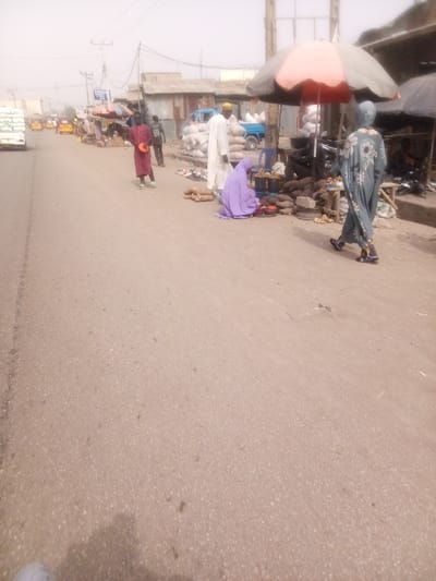 The image captures a bustling street scene, likely a market area, under what appears to be bright but hazy daylight. The setting suggests a developing urban or semi-urban environment, possibly in West Africa.
In the immediate foreground, a paved road, possibly asphalt, stretches across the bottom of the frame, showing some minor wear and debris. A faint shadow, likely from the photographer, is cast on the lower left corner of the road.
On the left side of the road, in the midground, a white cargo truck or van is seen moving away from the viewer. Further down the road, several yellow auto-rickshaws or small taxis are visible, indicating active transportation. Along this side of the street, there are indistinct stalls and buildings. A blue sign with white text, partially obscured by distance and haze, reads "ZARKAR" or "TARKAR" with "GENERAL STORE" below it. Overhead, power lines crisscross against the hazy sky.
The right side of the road is where most of the activity is concentrated, featuring a makeshift market. Several people are present:
* A person, possibly a child or young adult, in a red long-sleeved shirt and light-colored trousers, stands on the edge of the road, facing slightly left, holding a small orange bowl or container.
* To their right, a man in a long white traditional robe and a light yellow cap stands near a stack of large white sacks, presumably filled with goods like grain or produce. He is looking towards the market stalls.
* Further right, a person dressed in a full purple garment (like an abaya) is seated on the ground, attending to a small display of various items, including some red and light-colored produce, laid out in front of them. These items appear to be fruits or vegetables.
* A large red and white striped umbrella provides shade over another part of the market, where more goods are displayed. Underneath the umbrella, large, dark, cylindrical items, resembling yams or cassava, are stacked on the ground and on a simple wooden stand.
* On the far right, a person wearing a long, loose-fitting, light blue/grey robe with a patterned design and a head covering is walking away from the camera, along the side of the road.
Behind the market stalls, simple structures with corrugated metal roofs and walls line the street, consistent with informal market setups or residential buildings in such areas. A blue pickup truck, also loaded with white sacks, is parked behind the market activity.
The background is indistinct due to the haze, with vague shapes of distant buildings and power lines blending into the light, almost white, sky. The overall atmosphere is one of everyday commerce and movement in a sunlit, dusty environment.