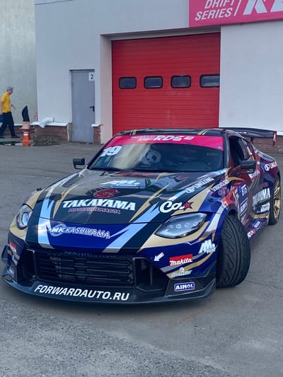 A custom-painted Nissan Z car, adorned with numerous sponsor logos and the number "19," is parked on a paved surface in front of a building with a large red industrial door. The car features a vibrant, multi-colored livery with metallic gold, blue, and white accents. Prominently displayed on the car's hood are the logos "TAKAYAMA" and "MK KASHIYYAMA." The front bumper has the website "FORWARDAUTO.RU" written in white. The car is angled, with its front wheels turned. To the left of the car, a person wearing a yellow shirt and a gray cap is walking away from the viewer. The building appears to be a garage or workshop, with a smaller gray door labeled "2" to the left of the red door. The ground is a rough, gray asphalt or concrete surface. A pink banner with "DRIFT SERIES" is visible at the top of the building. The scene is likely at a drift racing event or a related venue.