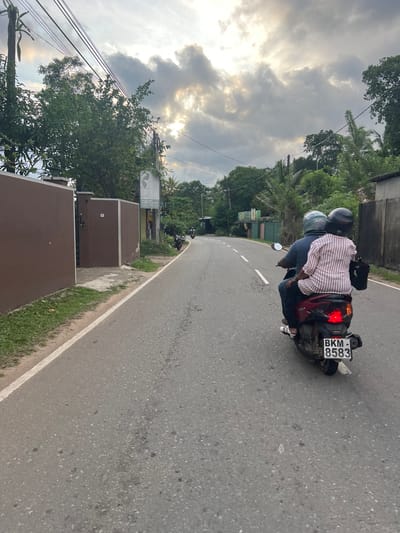 Angulugaha Junction, Sri Lanka: An eye-level, slightly rotated image depicts two people on a moped traveling on a gray asphalt road. The driver wears a gray helmet and a dark blue shirt. The passenger behind wears a red and white striped shirt. The moped's license plate reads "BKM-8583." The sky is cloudy, with the sun peeking through. The road is bordered by green vegetation on the left and a brown-painted wall. Trees and utility poles are visible in the background.