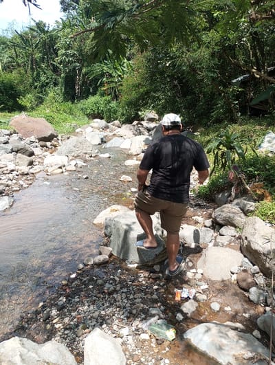 The scene depicts a male individual navigating a rocky riverbed in Cibeureum, Indonesia, under clear daylight conditions. The man, viewed from behind, wears a black t-shirt, brown cargo shorts, and blue slide sandals, with a white baseball cap worn backward. He is actively stepping from a smaller rock onto a larger, grey boulder on the right side of the frame, with his right foot planted and his left foot in mid-air or resting on the initial rock.
A shallow, clear-to-turbid river or stream flows from the left foreground towards the right, occupying the lower-left quadrant of the image. The riverbed and banks are densely covered with an assortment of grey, brown, and white rocks and pebbles of varying sizes. Visible on the rocky bank near the man's feet is scattered refuse, including a green plastic bag, an orange-labeled wrapper, and other unidentifiable plastic debris.
The background features dense, vibrant green tropical vegetation, including large broadleaf plants, possibly banana trees, and various shrubs, indicating a lush, overgrown environment. A faint structure, possibly a lean-to or small shack, is partially visible among the foliage on the far right. Sunlight illuminates the scene, casting shadows consistent with overhead sun.