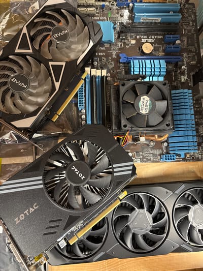 This image depicts computer hardware components, specifically several graphics cards and a motherboard, arranged on a wooden surface. The graphics cards are black with prominent cooling fans, and some have branding visible. One card clearly displays the "ZOTAC" logo, while another has a "2vfa" logo. The motherboard is a dark brown color with blue RAM slots and black heatsinks. It appears to be an ASUS brand motherboard, indicated by the "ASUS" logo and "MSA97 R2.0" text. The components are not in a functional computer system but are laid out as if for display or assembly. There is no indication of any specific location in Mülheim an der Ruhr, Germany, or any people present. The environment is a close-up shot of the computer parts, with a shallow depth of field that blurs the background.