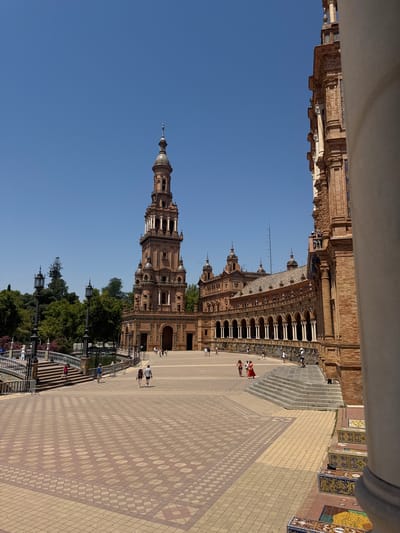 The image shows a bright, sunny day with a clear blue sky. The scene is an outdoor plaza with intricate architecture and tiled pathways. In the foreground, a wide expanse of light-colored tiles creates a patterned surface. Several people are visible walking on the plaza.
Dominating the background is a grand, ornate building with multiple towers and arches, constructed from reddish-brown brick. The building features extensive decorative elements, including columns and what appear to be tiled roofs. A tall, slender tower with a dome-like structure rises prominently from the building.
To the left of the building, a section of what seems to be a decorative railing or balustrade is visible, with trees and lampposts behind it. Further to the left, a part of another building, possibly a colonnade or walkway, is seen, also made of brick and featuring decorative stonework and white columns.
The image captures a lively public space, with people engaging in leisurely activities like walking. The overall atmosphere appears relaxed and pleasant, indicative of a tourist destination. The architecture suggests a historical or culturally significant location. Based on the provided location context, this is likely Plaza de España in Seville, Spain, known for its unique architecture and sprawling plaza.