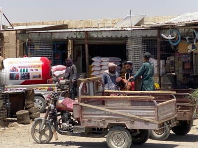 The scene is a street market in Panjwai, Afghanistan (latitude: 31.617, longitude: 65.599). Several men are standing around a truck with a red and white propane tank on the back, labeled in Farsi.  A man in a dark jacket stands near the truck, holding his hands in his pockets. Another man with a beard stands in front of the truck. He is looking to the side, and it's unclear if he is talking with another man standing in the back of a flatbed truck. This man is dressed in a gray outfit with a white head wrap, and he is looking towards the camera. The flatbed truck is loaded with a variety of objects, including a motorcycle and some loose items. There is a red motorcycle parked in the front of the truck.  A man dressed in green clothing is standing near the flatbed truck. He is facing the camera, and he appears to be talking to someone in the back of the truck, but only his back is visible. The street is dusty, and there are a few cinder blocks along the curb. There are multiple buildings with awnings in the background. The buildings are made of concrete and painted in a light beige color.  Several signs are visible, and all the text is written in the Farsi language. The scene is lit by sunlight.