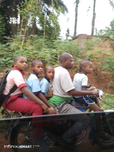 A man drives a motorcycle with four children riding with him, likely on their way to or from school. The children are wearing school uniforms; the first child on the back wears a maroon and cream uniform and a backpack, while the other children wear blue. The man wears a white shirt and dark pants. They are on a road surrounded by lush green trees and vegetation. The photograph has an "Infinix SMART 7 PLUS" watermark.