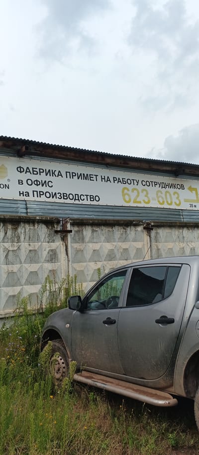 In Pirogovo, Russia, a gray pickup truck is parked amidst tall, green grass and weeds. The truck is positioned on the right side of the frame, with its front partially obscured. Behind the truck, a rough, textured concrete wall serves as a backdrop. Mounted on a corrugated metal structure above the wall is a large, white sign with black and yellow text. The sign reads in Russian: "FABRIKA PRIMET NA RABOTU SOTRUDNIKOV V OFIS na PROIZVODSTVO" which translates to "FACTORY ACCEPTS STAFF FOR WORK IN THE OFFICE for PRODUCTION". A phone number, "623-603", is prominently displayed in yellow on the right side of the sign, with an arrow pointing to the right and the text "35 M" below it. The sky above is overcast with gray clouds.