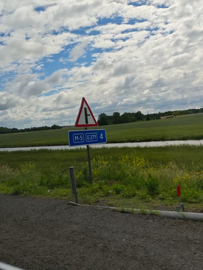 The image shows a road sign indicating a T-junction ahead, with an additional sign below displaying "M-5 E 271 4". The signs are positioned on a roadside with a gravel foreground and a grassy embankment with yellow wildflowers. In the background, there is a body of water, a green field, and a line of trees against a cloudy sky. The location is identified as Малевіцкі сельскі Савет, Belarus. There are no people or specific actions depicted.