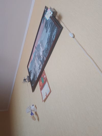 The image depicts an interior wall, likely in a residential setting. The wall has a light beige, textured wallpaper. A framed picture, featuring a mountain landscape with a castle or fortress, hangs on the wall. Below the picture, a small, decorative paper ornament, resembling a fan or a flower, is suspended by a string. To the right of the framed picture, a thin white electrical cord extends from the upper right corner of the frame, with a white inline switch located on the cord. Below the picture and to its left, a small, metallic lamp fixture is attached to the wall, with a visible light bulb. There is also a small, folded piece of paper or card attached to the wall near the bottom of the framed picture, featuring some text and graphics. The overall impression is of a decorated wall in a room. There is no indication of the specific location within the city of Bilhorod-Dnistrovskyi, Ukraine. There are no people present in the image.