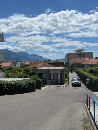 The image depicts a street scene in Bijela, Montenegro, under a partly cloudy blue sky. In the foreground, a paved road curves gently to the right, bordered by manicured green hedges and a blue metal railing on the right side. A man wearing a white t-shirt and dark shorts walks away from the camera on the right side of the road. A dark-colored sedan is driving up the street towards the camera. In the middle ground, several residential buildings with red-tiled roofs are visible, interspersed with lush green trees. A multi-story building with a white top structure stands prominently on the right. The background features a body of water, likely the Bay of Kotor, and a range of distant, mist-shrouded mountains. The scene appears to be a quiet, residential area on a bright, sunny day.