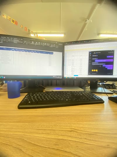 The image shows a desk with two computer monitors, a keyboard, and a blue object (possibly a cup or small container). The monitors display spreadsheets and a messaging application with text in Russian. The desk has a wood grain surface. The image is taken from a slightly elevated angle.

There is no information in the image to indicate the location is Luzhitsy, Russia.