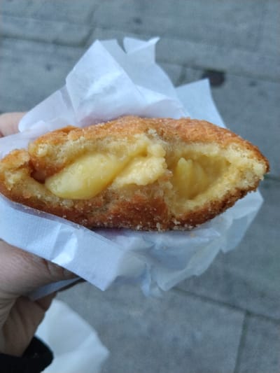A golden-brown, fried pastry is held in a white paper wrapper, sliced open to reveal a creamy, yellow filling. The crust appears to be sprinkled with a light coating of sugar. The texture of the pastry seems crispy. The filling is thick and smooth. The background shows a blurred grey surface, presumably pavement. A portion of a hand is visible holding the treat.