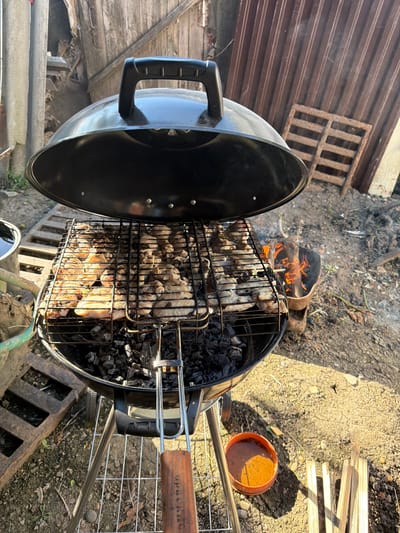 This media file captures an outdoor scene where food is being cooked over a charcoal grill. The primary objects are a black kettle-style grill with its lid open, revealing a metal grate laden with pieces of meat, likely chicken, being cooked over glowing charcoal. Fire can be seen burning in the background within a portion of the grill. The scene is set outdoors, with the ground appearing to be dirt and gravel, interspersed with some dry leaves and twigs. In the background, there are various discarded items including pieces of wood, a rusty metal grate placed on a corrugated metal surface, and some construction debris. To the right of the grill, a small orange bowl containing a dark liquid is visible, and further to the right, a stack of thin wooden planks rests on the ground. The lighting suggests it is daytime, with strong sunlight casting shadows. The overall impression is one of a casual, perhaps slightly rustic, outdoor cooking session. There is some visible text on the wooden handle of one of the grilling tools, appearing to be in Cyrillic script.