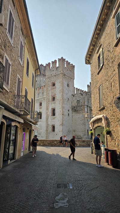 The image depicts a cobblestone street in Sirmione, Italy, leading towards the Scaligero Castle. On the left, a yellow building features shops with awnings and balconies. On the right, a stone building has an arched entrance and planters. Several people are present: two men are walking down the street, one in a dark t-shirt and shorts, the other in a dark polo shirt and shorts. A third man in a dark shirt and shorts is seen walking further down the street, and another man in a blue shirt and shorts is standing near trash bins. In the background, near the castle, a group of people are standing. The Scaligero Castle, a well-preserved medieval fortress, dominates the background with its stone towers and battlements under a clear sky. The lighting suggests it is daytime, with shadows cast on the cobblestone.