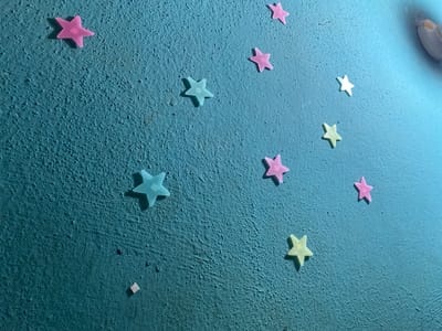 The image depicts a textured, painted blue wall adorned with numerous star-shaped stickers. The stickers vary in color, including pink, blue, and yellow. The wall has an uneven surface, and there is a slight color variation in the paint, with the right side appearing lighter. There are no people or discernible subjects present. The location information (Sabo, Nigeria) does not appear to be visually represented in the image.