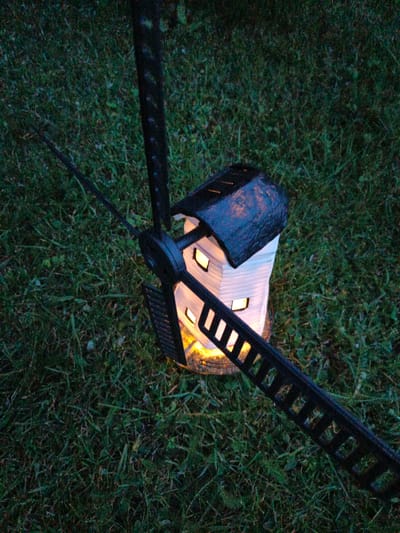 An illuminated windmill-shaped lantern sits on a patch of grass. The lantern is white with a dark, textured roof. It has several small, rectangular windows that emit a warm, yellow light. A black, three-pronged propeller mechanism extends from the side of the lantern, with one prong angled towards the foreground. The background is dark green grass. There are no people visible in the image.
