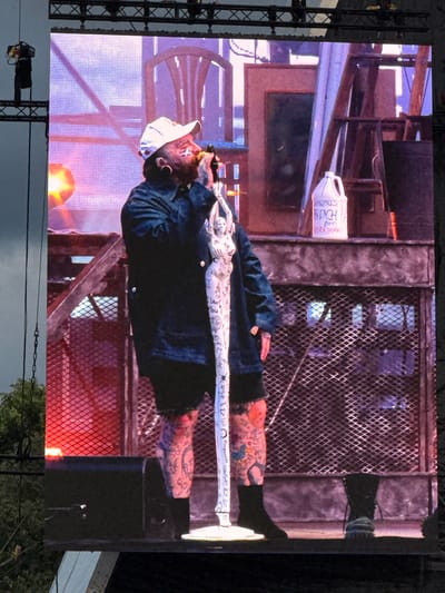 The image shows a large projection screen displaying a full-body shot of a male performing artist singing into a microphone. He is wearing a white baseball cap, a dark jacket, black shorts, and black socks. He has tattoos on his face, arms, and legs. The background on the screen features a rustic, industrial-themed stage set with various objects, including a ladder, a door, and a jug labeled "DEFINITELY BLEACH (NOT EVER DRINK)". The performer is standing on a stage, with speakers visible at the bottom. The screen is erected outdoors, with a grey sky and trees visible in the background to the left, suggesting it's a concert or festival. There is no indication of the specific location in Glasgow, United Kingdom.
