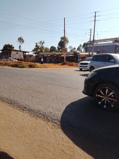 The image captures a street scene in Karingani ward, Kenya, under clear daylight. A paved asphalt road with an adjacent dirt shoulder occupies the foreground and midground. On the far right, the front right quarter of a black vehicle, featuring a tire with silver and black multi-spoke rims, is partially visible, casting a prominent shadow onto the road. In the midground, a silver SUV (license plate KCL 486L) and a white SUV are positioned on the road. Along the left side of the paved area, a row of informal commercial structures with corrugated iron roofs and various signs is present. One individual, dressed in white, is observed walking left away from these structures. To the far left, a sign reading "Treedom Gardens" stands near wooden framework structures, behind which a white sedan is parked. Multiple utility poles supporting numerous overhead power lines punctuate the scene. In the background, a multi-story building is visible to the right, beyond which scattered trees and vegetation extend under a clear blue sky.