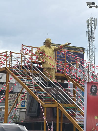 A golden statue of a man is positioned on a yellow metal platform with stairs, adorned with small red and white flags. The statue is wearing traditional Indian attire. Behind the statue, a large banner advertises "Malla Reddy Engineering Aspirants" and "Malla Reddy Institute of Engineering and Technology," featuring contact information and course details. A cell tower and a security camera are visible in the background against an overcast sky. To the right of the statue, a red banner displays a portrait of Che Guevara. The scene appears to be in Kurnool, India, as indicated by text in Telugu script on signs in the background, including "మదర్స" and "భోజనం." The overall atmosphere is one of an outdoor urban setting, possibly a public space or educational institution campus, decorated for an event.