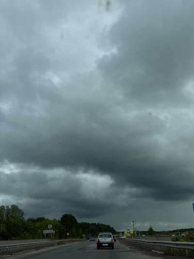 The image shows a view from a roadside in Совхозный, Russia, on a cloudy day. A gray car is driving on the road, which is flanked by a guardrail and trees on the right side. Numerous road signs are visible along the roadside. The sky is filled with heavy, dark clouds, suggesting an overcast or stormy weather condition.