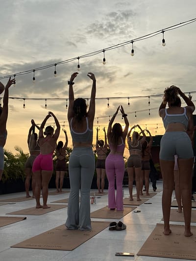 The image shows a group of women practicing yoga outside, likely in Tibubeneng, Indonesia. They are standing on yoga mats, facing away from the viewer, with their arms raised above their heads. The women are wearing athletic clothing in various colors. Overhead string lights are visible, and the sky is a hazy yellow, indicating a sunset or sunrise. In the background, palm trees are visible. A pair of shoes and a phone are lying to the side of the mat.