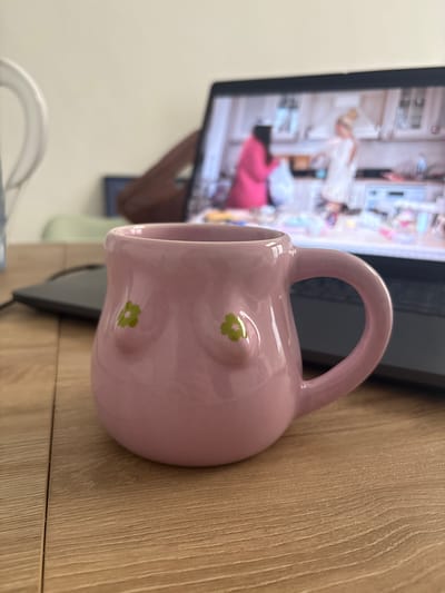 A light pink ceramic mug with green flower designs is positioned on a light brown wooden surface. The mug has a large handle. Behind the mug, there's a black laptop with an image on the screen depicting a person in what seems to be a room with various objects. Part of a white plastic object is visible in the lower-left corner of the image. The scene is indoors. There's no direct indication that this is in Turin, Italy.