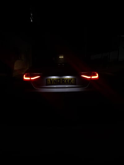A white car is parked at night, its brake lights illuminated. The car is facing away from the camera, and a yellow license plate with the registration "VN12RXK" is visible. The background is dark, with some faint light sources suggesting a residential area. The scene appears to be in Dolni Tsibar, Bulgaria. No people are visible in the image.