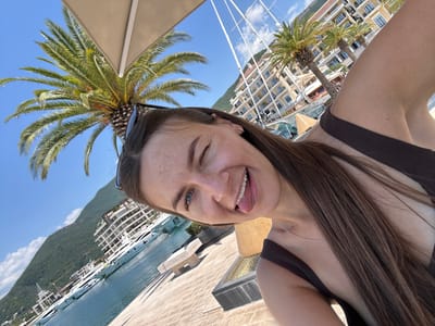 A woman is taking a selfie in Donja Lastva, Montenegro. She is wearing a brown bikini top and a silver necklace. Her eyes are blue and she is winking. She has long brown hair and freckles on her face. She is sticking her tongue out at the camera. In the background, there are palm trees, a body of water with several yachts, buildings, and mountains. The sky is blue and there are some clouds. The woman is standing on a paved area.