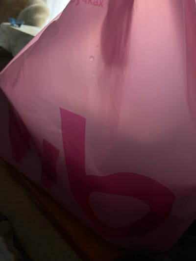 A close-up shot captures a pink bag with a large, dark pink "C" shape emblazoned on its side. The bag's surface is slightly crinkled and shows some subtle light reflections and faint water spots. The texture appears to be a smooth, slightly shiny material, possibly plastic or coated paper.
In the lower left corner, a portion of white material, possibly another bag or packaging, is visible, with a small blue clip attached. Faint Cyrillic text, "ручках" (which translates to "handles" in English), is printed vertically on the pink bag to the left of the "C" shape.
The lighting suggests an indoor setting, possibly dimly lit. The background is dark and out of focus, with hints of what might be a wooden surface or other objects. The overall impression is a snapshot of an object, likely a shopping bag, in a casual, everyday context. The location context is identified as Minyar, Russia. No people are visible, and the activity is simply the presentation of the object.