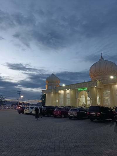 This image depicts a mosque, identified by the text "BAITUL HUDA" displayed prominently in green neon letters above the entrance. The mosque features classic Islamic architecture with a large dome, a smaller dome, and decorative minaret-like structures. The building is illuminated, suggesting it's either dusk or dawn. The sky is overcast with dramatic, dark clouds, creating a moody atmosphere.
In front of the mosque, a paved parking lot is filled with various cars, including SUVs and sedans. A few individuals are visible in the parking lot and near the mosque entrance, some appearing to be walking or standing. One person in the foreground is wearing dark clothing and a headscarf, standing in the middle of the parking area.
The scene is set in a location that appears to be a public space, possibly outside of a town or city, given the open sky and distant lights. The overall mood is serene, with the illuminated mosque standing out against the darkening sky. The time of day appears to be early evening, as indicated by the dim natural light and the artificial lighting of the mosque and streetlights. The weather seems cloudy and possibly humid, typical of tropical regions. The presence of cars suggests it is a place of gathering or worship, with people arriving or departing.