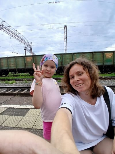 A selfie is taken on a cloudy day at a train station in Kropachyovo, Russia. The photographer, an adult woman with curly brown hair, is smiling and wearing a white t-shirt. To her left, a young girl with a patterned headscarf and a pink t-shirt is making a peace sign with her fingers. Behind them, a green freight train is visible, with overhead power lines and a network of tracks in the foreground. The ground is paved with textured tiles.