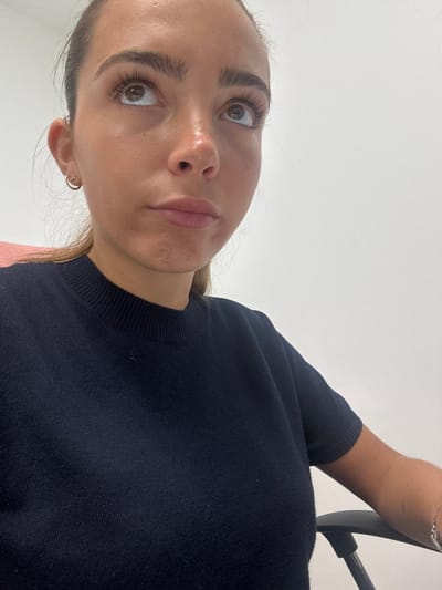 The image depicts a woman looking upward at the camera. She has fair skin, brown eyes, and dark eyebrows. She is wearing a dark blue knit top. Her hair appears to be pulled back, and she is wearing small hoop earrings. The background is a plain white wall. The location is not discernible from the provided image.