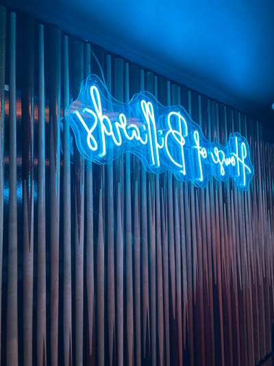 This image depicts the interior of the House of Billiards located at Varkensstraat 2, 6811 GM Arnhem, Netherlands. The central subject is a vibrant blue neon sign, horizontally reversed, displaying the cursive text "House of Billiards." The sign is mounted on a wall decorated with numerous vertically oriented wooden rods, which strongly resemble billiard cues, creating a textured background. The blue light from the neon sign illuminates the immediate area, casting a cool blue hue across the wall and the plain, dark blue ceiling visible in the upper portion of the frame. No individuals, specific actions, or interactions are visible in the scene. The overall environment suggests an entertainment venue or bar.