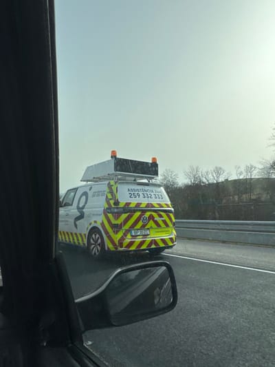 An image captured from the interior of a moving vehicle on a highway near Macedo de Cavaleiros, Portugal, depicts the rear-three-quarter view of a white Volkswagen ID. Buzz Cargo van ahead. The van features a large "G" logo on its side and the text "Assistência 24H" (24-hour assistance) with the phone number "259 332 333" on both its side and rear. The rear and lower sides of the van are covered in reflective red and yellow/lime green chevron markings. The vehicle's license plate, "BP 30 ZC," is clearly visible, with the word "transmontano" printed below it. Mounted on the van's roof is a dark, rectangular structure, possibly a portable warning sign or message panel, equipped with two amber strobe lights at its upper corners. The road surface is dark asphalt and appears wet. To the right of the van, a grey concrete or metal guardrail separates the highway from a natural embankment with bare deciduous trees and distant hills under an overcast sky. The foreground of the image shows the black interior A-pillar and a portion of the driver's side mirror of the observer's vehicle. Both vehicles appear to be in motion, traveling in the same direction.