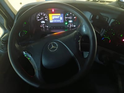 This media file captures the interior of a vehicle's cabin, focusing on the steering wheel and dashboard. The scene is illuminated by the vehicle's internal lighting, suggesting it might be nighttime or a dimly lit environment.
The primary objects are a black steering wheel with integrated controls and a Mercedes-Benz logo at its center, and a dashboard featuring multiple gauges and a digital display screen. The gauges include a speedometer on the left, a tachometer on the right, and smaller gauges below them. The digital screen displays a yellow warning message with Cyrillic text. The text appears to be a system message, possibly related to a vehicle status or alert.
Visible text on the dashboard includes:
- "СТОЯНОЧНЫЙ ТОРМОЗ" (Parking brake)
- "ВЫКЛЮЧЕН" (Disengaged)
- "ЗАМЕНА МАСЛА БЛИЖАЙШИЙ" (Oil change next)
Green indicator lights are visible on the steering wheel controls and the dashboard. A red light is also illuminated on the dashboard, likely indicating a warning or alert. The overall impression is one of a functional vehicle cabin, possibly a truck or a commercial vehicle, given the size and layout of the dashboard. The image is taken from the driver's perspective, looking forward. The location context suggests the image was taken in Қопа ауылдық округі, Kazakhstan.