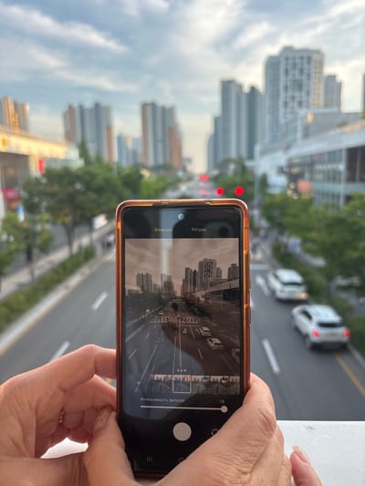 The media file shows a close-up of a person's hands holding a smartphone. The phone's screen displays a photo of a city skyline, with tall buildings and a road with cars in the foreground. The phone is oriented vertically, and the image on the screen is also vertical. The camera is positioned at an angle that suggests it's being used to take a photo or video of the cityscape outside. The background of the scene shows a blurred view of buildings and trees, indicating an outdoor urban environment. The sky appears overcast, suggesting it might be daytime or late afternoon.

Visible text on the phone screen includes "Ретушь" (Retouch) and "Фильтры" (Filters) in Russian, suggesting the phone's user interface is in that language. There are also options like "B & W" (Black & White) and "Интенсивность фильтра" (Filter Intensity) visible, indicating photo editing features. The overall mood conveyed is one of capturing a moment or enhancing a visual, with the focus on the technology and the cityscape it's framing. The presence of hands holding the device points to a personal activity, likely photography or videography. The context provided places the scene in Incheon, South Korea.