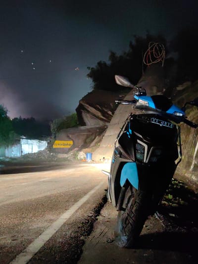 A dark, nighttime scene unfolds on a rural road near Kulang, India. The primary subject is a parked scooter, its front half illuminated by its own headlights, casting a bright beam forward. The scooter is black and blue, with a license plate that reads "HP 56A 4814". In the background, a steep, rocky hillside looms, partially obscured by darkness. A yellow sign with Hindi text is visible on the hillside. To the left, a corrugated metal structure and a pile of debris suggest construction or disarray. The sky is dark with a few distant lights, possibly lanterns or distant fires, creating a somewhat eerie atmosphere. The road surface is rough, with loose stones and gravel visible along the edge. The overall mood is quiet and isolated, with the scooter's headlights being the only strong source of light in the immediate vicinity.