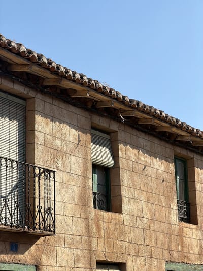The image shows a close-up view of the exterior of a building in Arcahueja, Spain. The building is constructed with large, tan-colored stone blocks. A tiled roof with an overhang is visible at the top of the image. Below the roof, there are two windows with green frames. One window is on the left side and has a white slatted blind. In front of this window is a black wrought-iron balcony. A blue tile with the number "3" is affixed to the wall below the balcony. The second window is on the right side and also has a green frame and a black wrought-iron grate. The sky above is clear and blue. The overall scene is static and captures a portion of a building's facade.