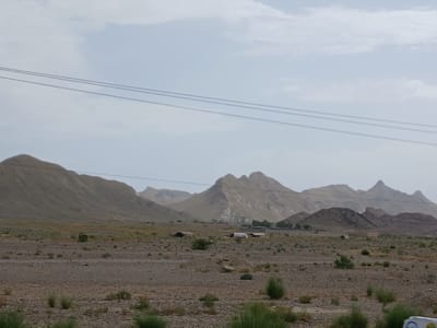 The image depicts a barren, mountainous landscape with sparse vegetation and a few structures. In the foreground, the ground is covered with gravel and small rocks, with patches of dry grass and shrubs. In the midground, several tents and simple buildings are scattered across the terrain. These structures appear to be temporary or basic dwellings. In the background, rolling hills and mountains dominate the horizon, with hazy peaks suggesting a dry, arid environment. Three power lines stretch horizontally across the upper portion of the image, indicating the presence of infrastructure. The sky is overcast with light clouds. The scene suggests a remote, sparsely populated area, consistent with the rural outskirts of Qilla Saifullah, Pakistan, known for its rugged terrain and dry climate. There are no visible people or animals in the frame.