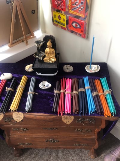 This is an indoor, eye-level, medium shot of a display of candles. The candles are arranged in bundles on a table covered with a purple velvet cloth. The table is placed in front of a wooden dresser with ornate drawer pulls. Behind the table, a golden Buddha statue sits on a black base with a water feature. To the left, an easel stands against a white wall. To the right, a portion of a colorful, abstract painting is visible on the wall. There are also various decorative stones and a small ceramic dish on the table. The lighting suggests natural light coming from a window. The scene appears to be a retail display, possibly in a shop or market stall. There are no people present in the image. The location is identified as Rother, United Kingdom.