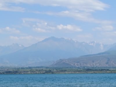 This image depicts a tranquil natural landscape. In the foreground, a body of water with a turquoise hue stretches across the frame, its surface gently rippled. Beyond the water, a verdant shoreline is visible, dotted with scattered trees and greenery. Further in the distance, a range of mountains rises, their peaks softened by atmospheric haze. The sky above is a pale blue, adorned with scattered white clouds. The overall scene conveys a sense of peacefulness and natural beauty. The city of Kirpichny, Kyrgyzstan is known for its mountainous terrain and proximity to water bodies like Issyk-Kul Lake, which this image could potentially represent. However, without further context or specific landmarks, it is impossible to definitively confirm the location.
