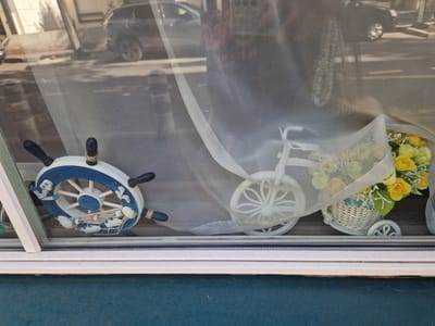The image shows a window display. On the left is a decorative ship's wheel with a nautical theme, featuring seagulls, shells, and fish. To the right of the ship's wheel is a small white decorative bicycle with a basket filled with yellow roses. The display is viewed through a window, and the background shows reflections of cars and the street outside, suggesting an urban setting. The scene is framed by the window and a ledge. The overall impression is one of decorative objects displayed behind glass. The location is the city of Antony, France.