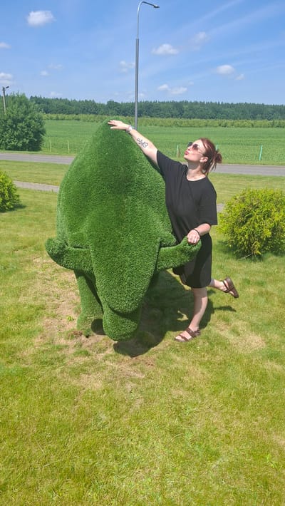 A woman with red hair pulled into a bun and wearing sunglasses and a black t-shirt and skirt poses with a large, green, topiary sculpture of an elephant. She is standing with her left hand on the elephant's head and her right arm extended upwards. Her right leg is bent and lifted behind her. The sculpture is made of artificial grass and is situated on a grassy lawn. In the background, there is a field of green crops, a line of trees, and a bright blue sky with scattered white clouds. A lamppost is visible behind the woman. The location is identified as Дзяржынскі сельскі Савет, Belarus.